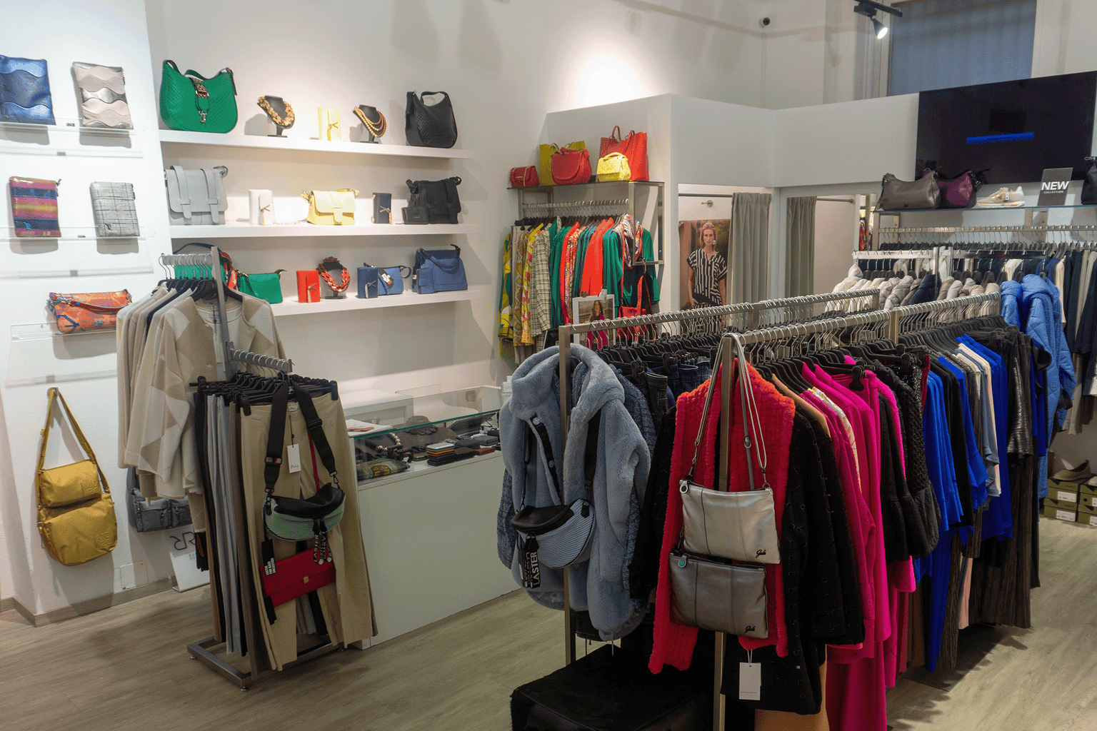 Joseph Ribkoff Flagship Store Wien Innenansicht mit farbenfroher Damenmode in Pink, Blau und Grün aus feinen Stoffen sowie Taschen aus Leder und Textil in verschiedenen Farben, präsentiert in eleganter Boutique Atmosphäre. Blick auf die Kassa