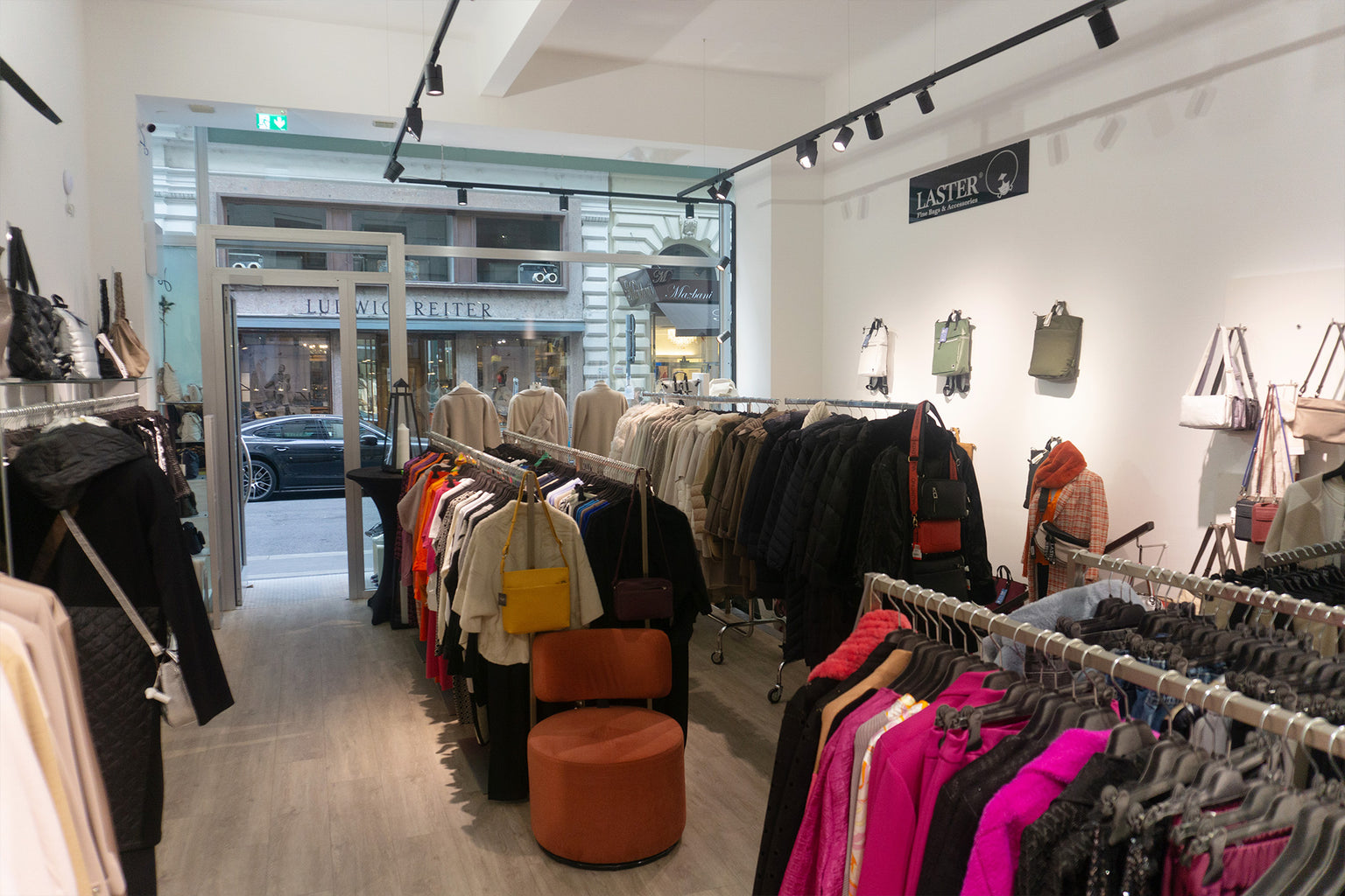 Innenansicht des Joseph Ribkoff Flagship Store Wien mit Blick auf den Eingang zur Führichgasse. Im Vordergrund Kleiderstangen mit farbenfroher Damenmode, darunter Blazer, Mäntel und Jacken in Pink, Rot, Beige, Schwarz und kräftigem Fuchsia aus hochwertigen Stoffen wie Wolle, Baumwolle und Mischgewebe. Auf der rechten Seite elegante Handtaschen aus Leder und Textil in neutralen Tönen wie Grau, Beige und Schwarz. Links Mäntel und Jacken in gedeckten Farben, kombiniert mit Taschen in Gelb und Rot. Der helle Bo