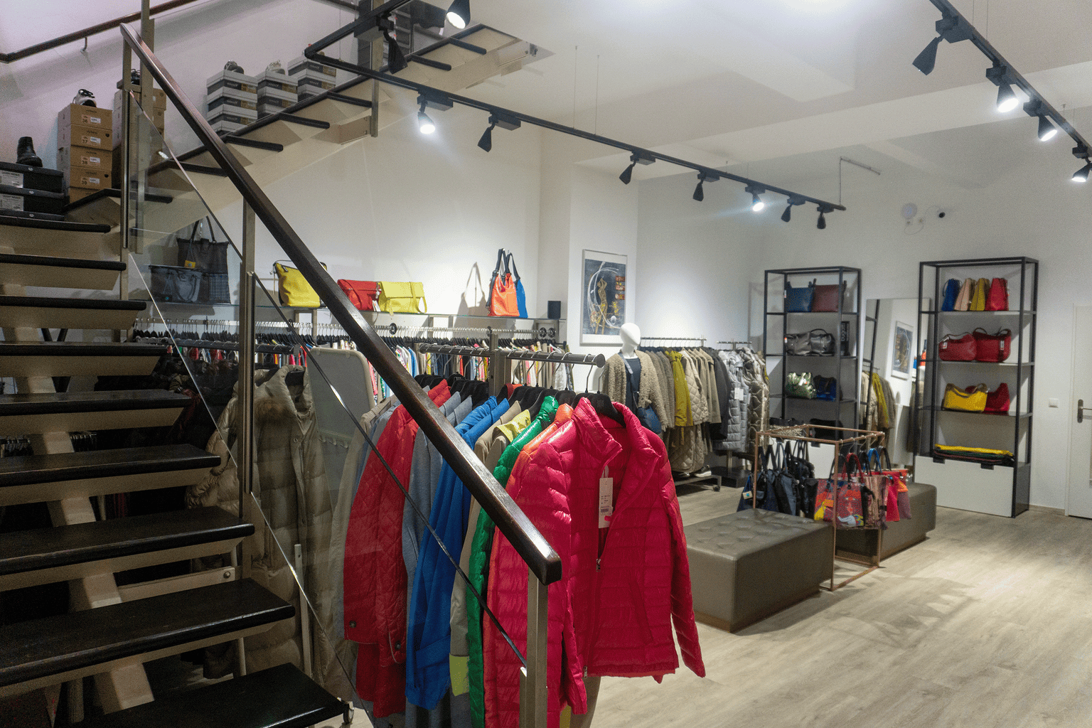 Innenansicht der unteren Etage im Joseph Ribkoff Flagship Store Wien mit Blick auf moderne Boutique Einrichtung. Im Vordergrund eine Kleiderstange mit Daunenjacken in kräftigen Farben wie Pink, Rot, Blau und Grün aus glänzendem Nylonmaterial. Daneben Mäntel und Jacken in Beige, Grau und Schwarz aus Wolle und Mischgewebe. Auf Regalen elegante Handtaschen aus Leder in Gelb, Rot, Grün und Schwarz sowie Shopper aus Textil mit bunten Prints. Links eine Treppe mit Glasgeländer, im Hintergrund Sitzgelegenheiten au