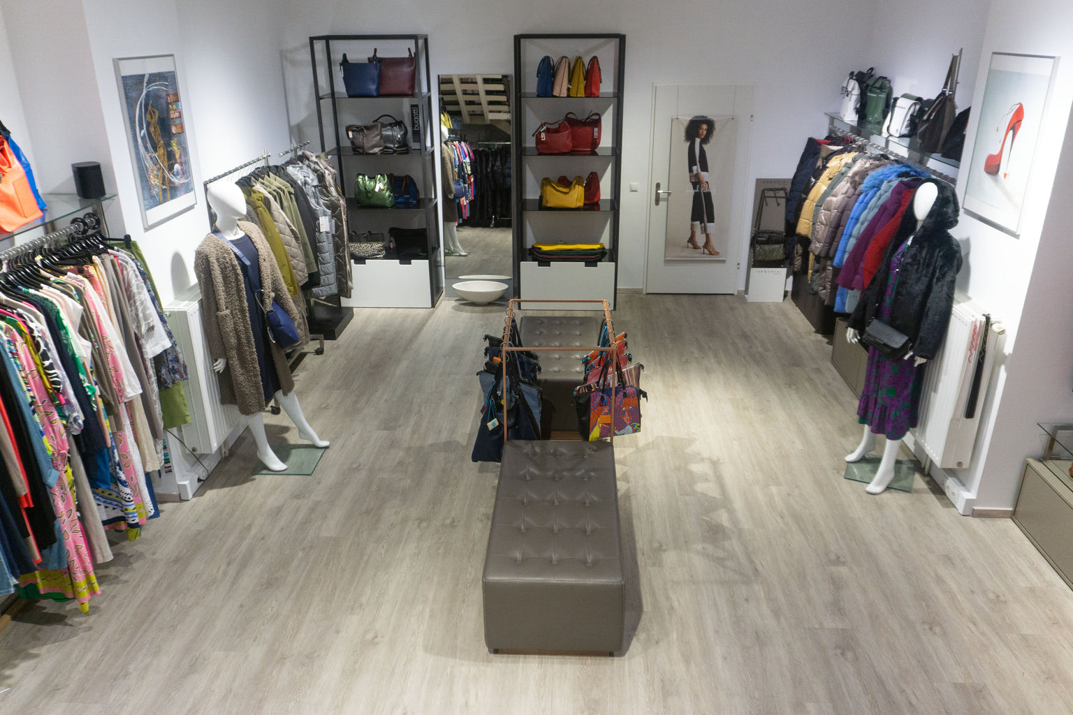 Innenansicht der unteren Etage des Joseph Ribkoff Flagship Store Wien mit Blick auf ein offenes Boutique Ambiente. Links eine Kollektion farbenfroher Kleider in Rosa, Grün, Blau und gemusterten Stoffen aus Viskose und Baumwolle. Rechts Mäntel und Jacken in gedeckten Farben wie Schwarz, Beige und Grau sowie kräftigen Tönen in Blau, Rot und Lila, gefertigt aus Wolle, Daunen und Mischgewebe. In der Mitte eine Sitzbank aus grauem Leder mit Taschenpräsentation, darunter Handtaschen und Shopper aus Leder und Text